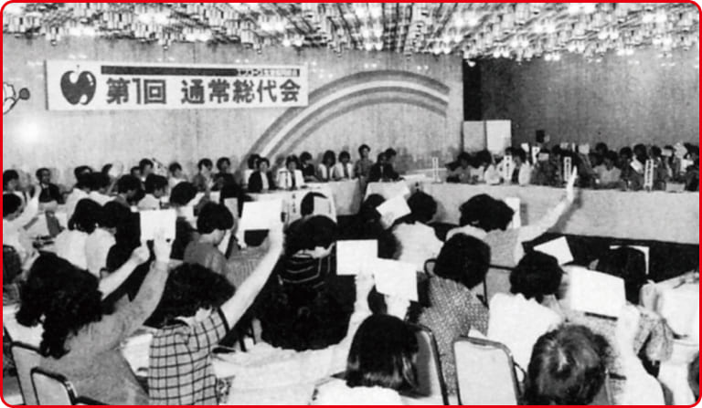 第1回通常総代会の写真。ホールに多くの人が集まり、中心を囲むように椅子に座っている。その中の数人が手を挙げている様子。